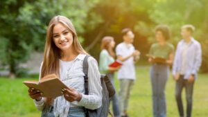 Young woman holding a book and smiling in a park, representing scholarship opportunities and academic success, with friends in the background.